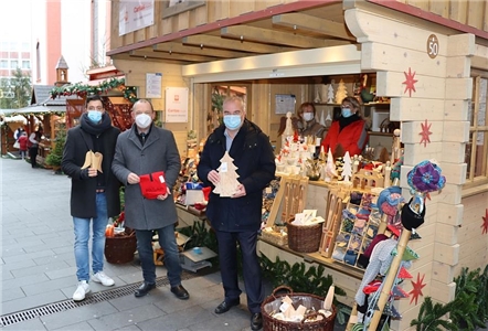 Die drei Personen stehen vor dem Weihnachtsmark und halten unterschiedliche Verkaufsartikel vor sich. 