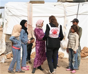 Eine Frau mit einem Caritas-Logo auf dem Rücken spricht vor einem Zelt mit Flüchtlingen. Die Zeltwände sind am Boden mit großen Steinen beschwert. Die Die geflohende Frau undhre Töchter tragen Kopftüc