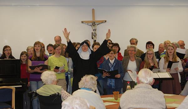 Vor einer breiten weißen Wand, an der in der Mitte ein Holzkreuz mit silbernen Korpus hängt, haben sich viele Sänger unterschiedlichen Alters aufgestellt. Sie halten Notenblätter in ihren Händen. Vor 