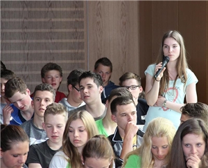 Wir blicken in die in die Reihen der Schüler. Rechts steht ein Schülerin. Sie spricht gerade in ein Mikrofon, welches sie in der rechten Hand hält.