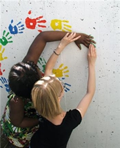 Ein Mädchen mit heller Hautfarbe führt die Hand eines dunkelhäutigen Mädchens und drück ihre Hand fest auf eine helle Wand. Im Halbkreis sind weiter bunte Hände auf diese  Wand gedruckt.