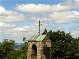 Unter einem blau-weißen Himmel werden Baumkronen sichtbar. Vor ihnen steht ein Turm mit einem grünen Kupferdach. Darauf ist ein schlichtes Kreuz angebracht.