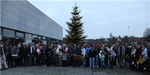 Vor einem großen hohen mit Lichtern geschmückten Weihnachtsbaum haben sich auf einem großen Platz in mehreren Reihen Flüchtlinge und Bewohner von Neuhof zu einer großen Gruppe aufgestellt. 