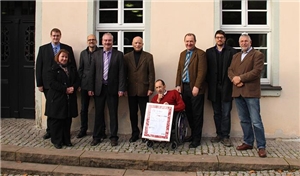 Die Beteiligten haben sich vor der Geschäftsstelle des Caritasverbandes in der Wilhelmstr. 2 in Fulda in einer Reihe aufgestellt. Herr Booelken sitzt in der Mitte des im Rollstuhl und hält die Urkunde