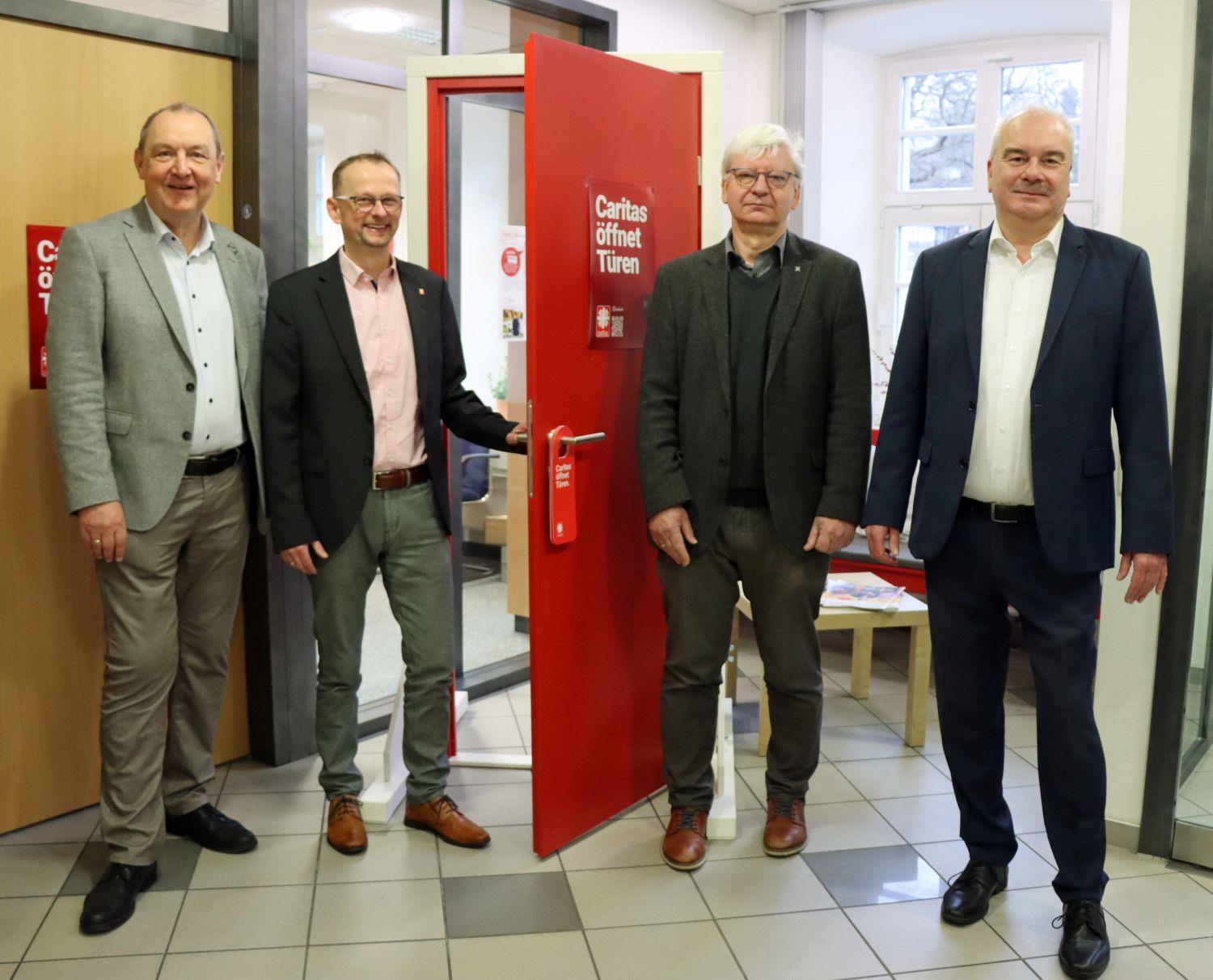 Um die rote Caritastür herum stehen Ansgar Erb von der Fuldaer Diözesan-Caritas, Rainer Broßmann und Ludwig Borowik von der Regional-Caritas MKK sowie Caritasdirektor Juch