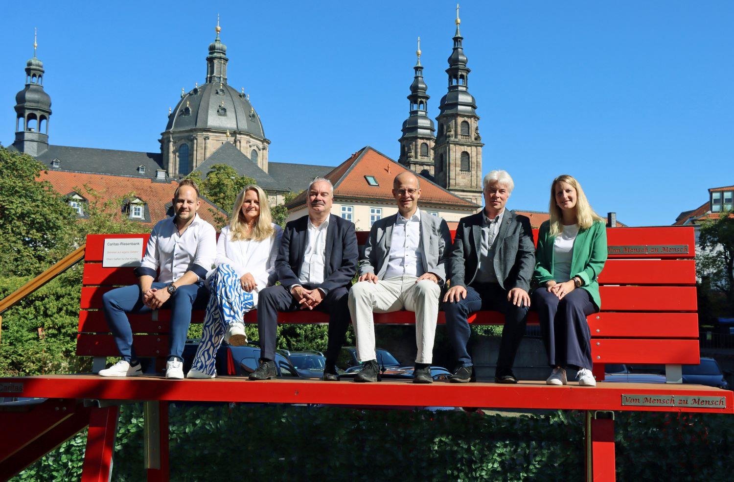 Auf der Riesenbank sitzen Alexander Ponelies, Susanne Saradj, Caritasdirektor Markus Juch, Michael Brand, Holger Franz sowie Ann-Katrin Jehn