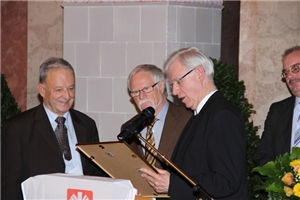 Professor Stanke hat Preisträger zum Pult geholt. Er blickt gemeinsam mit zwei der Preisträger auf die eingerahmte Urkunde.