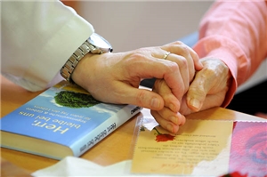 Die Hand einer Seniorin liegt neben einem Buch auf einer Karte. Eine zweite Hand fast über das Buch die Finger der Seniorin. 