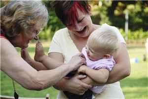 Eine ältere Dame reicht die Hand nach einem blonden Kleinkind, welches in den Armen einer jungen Frau liegt