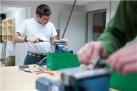 Wohnverbund_Haus_Agathaberg_QUANTUM Im Hintergrund bearbeitet ein Bewohner das Holz, im Vordergrund sind zwei Hände, die ein Stück Holz im Schraubstock einspannen