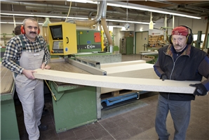 Auf dem Bild sind zwei Mitarbeiter die fertige Plattenzuschnitte von der Maschine wegtragen.