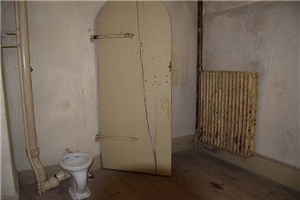 Wir sehen einen alten kahlen Kellerraum. Die Tür hat keine Klinke an der Innenseite. Ein Toilettenkörper ohne Garnitur ist neben der Tür gebaut.
