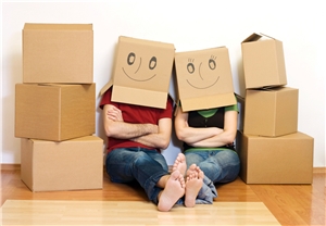 Zwei Menschen sitzen zwischen Umzugskisten auf dem Boden und tragen Kisten über dem Kopf, auf denen lächelnde Smileys aufgemalt sind