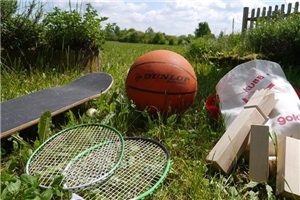 Auf einer grünen Wiese liegen verschiedene Speile bereit. Tennisschläger, ein Kubb-Spiel, ein Skateboard und ein Fußball laden zum Benutzen ein. 