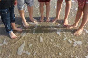 Kinder haben sich barfuß im Halbkreis am Strand aufgestellt. Zwischen Ihren Füßen rinnen leichte Wellen hindurch.
