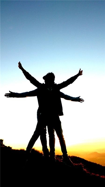 Zwei Jugendliche stehen bei einem Sonnenuntergang auf einem Berg. Sie haben sich so hingestellt, dass  ihre Silhouette einer Windmühle gleicht.