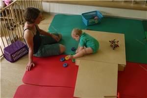 Kleinkindbetreuung Ein helles Zimmer ist mit grünen und roten Matten ausgelegt. Auf ihnen stehen Holzelemente, worauf ein Kleinkind sitzt. Eine Erzieherin schaut ihm zu.