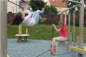 Wir befinden uns auf einem Spielplatz. Ein Mädchen klettert am waagerechten Seil  zur anderen Seite des Kletterturmes. Ein anderes Mädchen balanciert auf einem Seil.