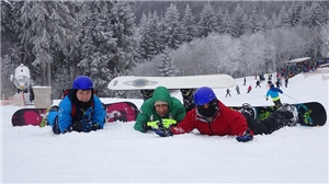 Drei Jugendliche liegen in Schneeanzügen und Helmen im Schnee.