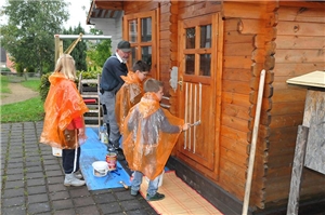 Eine Massivholzhütte wird von Kindern und Erwachsenen mit einer hellen Farbe gestrichen.