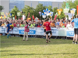 Staffelstabübergabe In derr bunten Wechselzone versuchen alle Teams mit großen, auffälligen Schildern auf sich aufmerksam zu machen. Ein Läufer der Caritas übergibt gerade den Staffelstab.