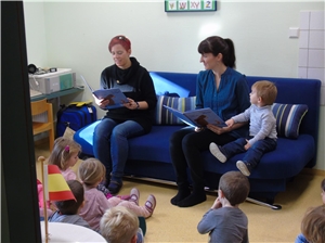 zwei Frauen sitzen mit Büchern auf einem Sofa, Kinder sitzen drum herum