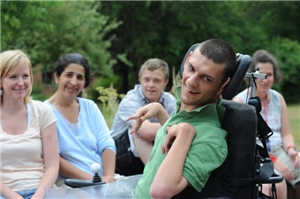Ein Mann mit Behinderung im Vordergrund, im Hintergrund sitzt eine Gruppe Menschen