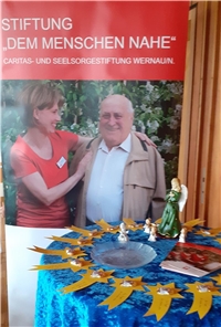 Ein Plakat steht hinter einem Tisch mit Engeln darauf. Es zeigt eine junge Frau und einen älteren Mann