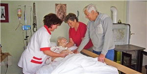 Pflegerin und Angehörige bei pflegebedürftiger Frau am Bett
