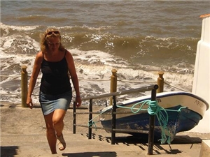Eine Frau in sommerlicher Kleidung steigt Stufen am Strand hinauf. Neben ihr liegt ein kleines Boot. Die Sonne scheint.