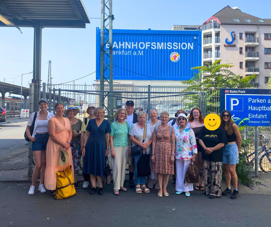 Eine Gruppe steht in Frankfurt vor einem Schild mit Logo der Bahnhofsmission Frankfurt