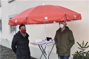 Unter einem Caritas-Schirm steht 2 Mitarbeiterinnen mit Büromaterial
