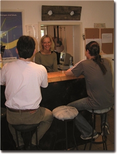 Zwei Männer sitzen auf Barhockern im Kontaktladen, lächelnde Mitarbeiterin