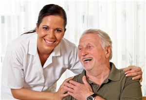 Fröhlich lachende Pflegerin steht neben sitzendem Senior