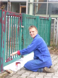Ein Jugendlicher kniet auf dem Boden und streicht einen Zaun mit Farbe.