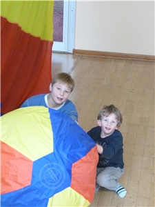 Zwei Jungen sitzen in der Turnhalle und halten die Enden eines bunten Fallschirms fest.