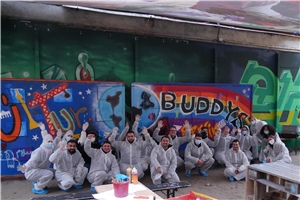 Viele junge Menschen die mit Schutzanzügen vor einer Graffiti-Wand stehen, auf der Kulturbuddys drauf steht