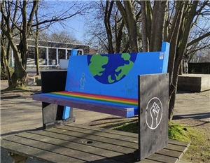 Eine hellblau bemalte Parkbank mit Weltkugel, Regenbogenfarben auf der Sitzfläche und Black Lives Matter Symbol an der Seite