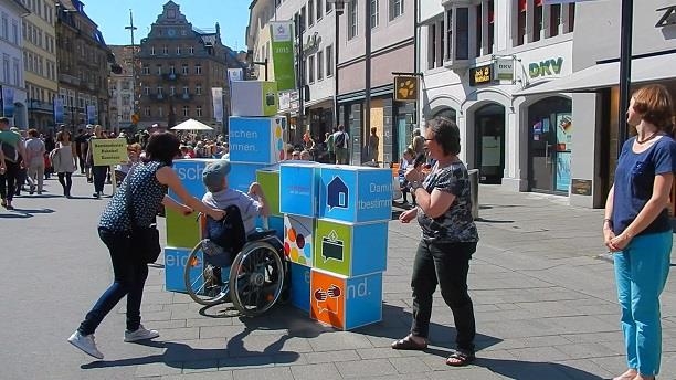 Würfelwand auf die ein Rollstuhlfahrer zufährt (Caritas Konstanz)