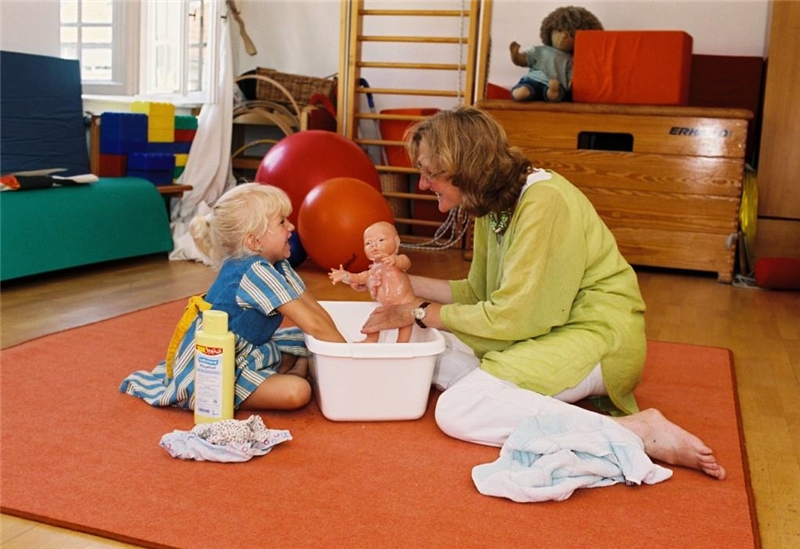 : Ein kleines Mädchen und eine Frau sitzen auf dem Boden, zwischen ihnen eine Baby-Badewanne. Das Mädchen hält die Arme in die Wanne, die Frau hält eine Puppe in der Hand.