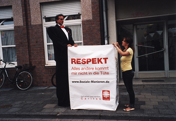 Ein Mann im Anzug (der auf Stelzen steht) und eine Frau, die neben einer übergroßen Respekttüte stehen, die sich auf einem Bürgersteig befindette stehen (Caritas)