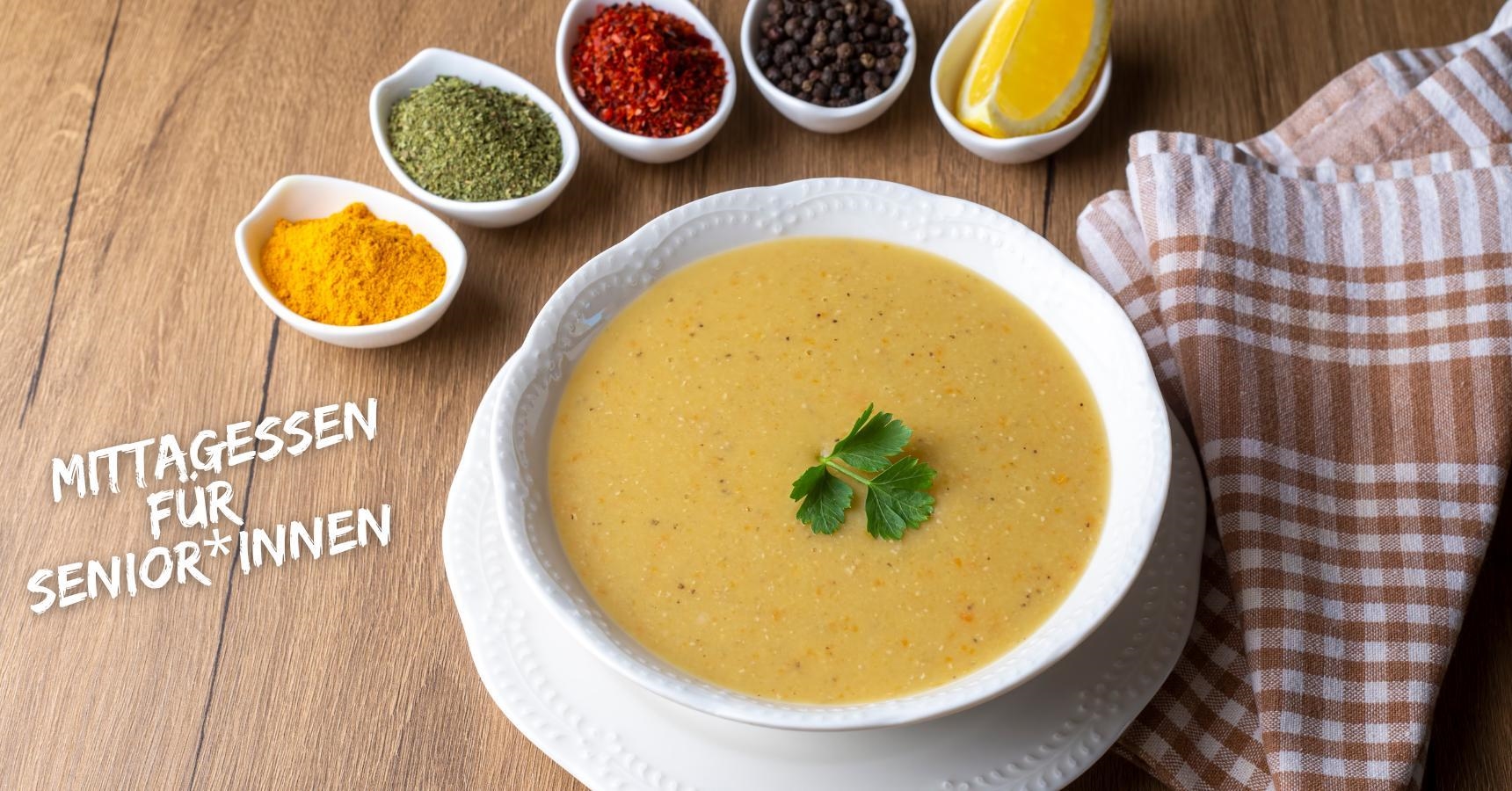 Ein Teller mit gelblicher Linsensuppe; rund um den Teller stehen kleine Schälchen mit Gewürzen und einem Viertel Zitrone. Rechts liegt ein braun kariertes Küchenhandtuch.