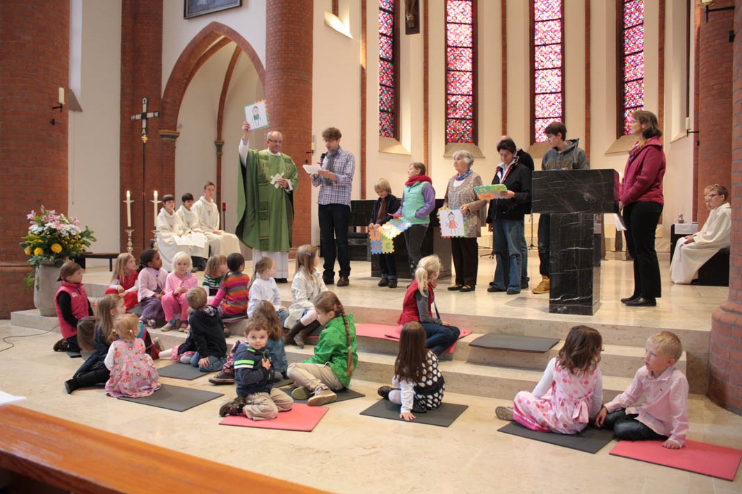 Kinder sitzen auf den Altarstufen der Kirche Herz-Jesu Lübeck und schauen auf den Pfarrer, der ein gemaltes Kinderbild erläutert, das eine Person zeigt. (Manuela Siegerink-Schröder)