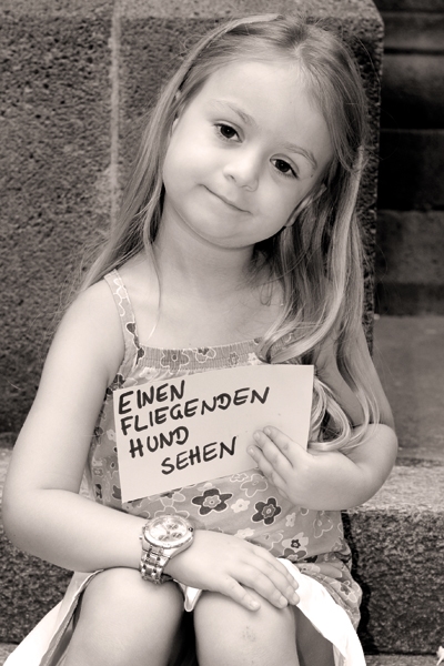 Josephine (5) auf dem Brüsseler Platz in Köln. Er hält ein Schild mit der Aufschrift "Einen fliegenden Hund sehen". Die Aufnahme ist in schwarz-weiß. (Tanja Anlauf (Fotoprojekt „Unsere Zukunft“)