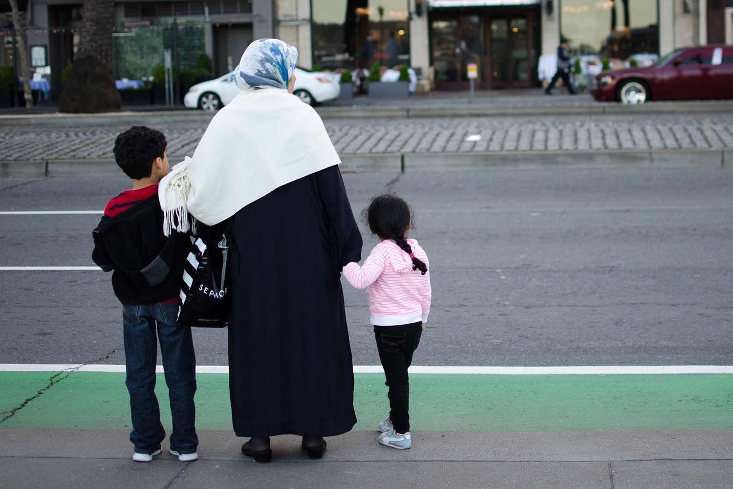 Eine Frau mit Kopftuch und jeweils einem Kind an der Hand steht auf einem Bürgersteig.