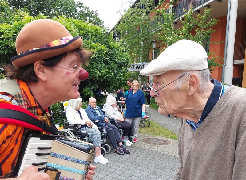 Ein verkliedeter Clowndoktor und ein Bewohner des Altenheims neigen die Köpfe zueinander und sehen sich direkt in die Augen. Beide sind von der Seite abgebildet.