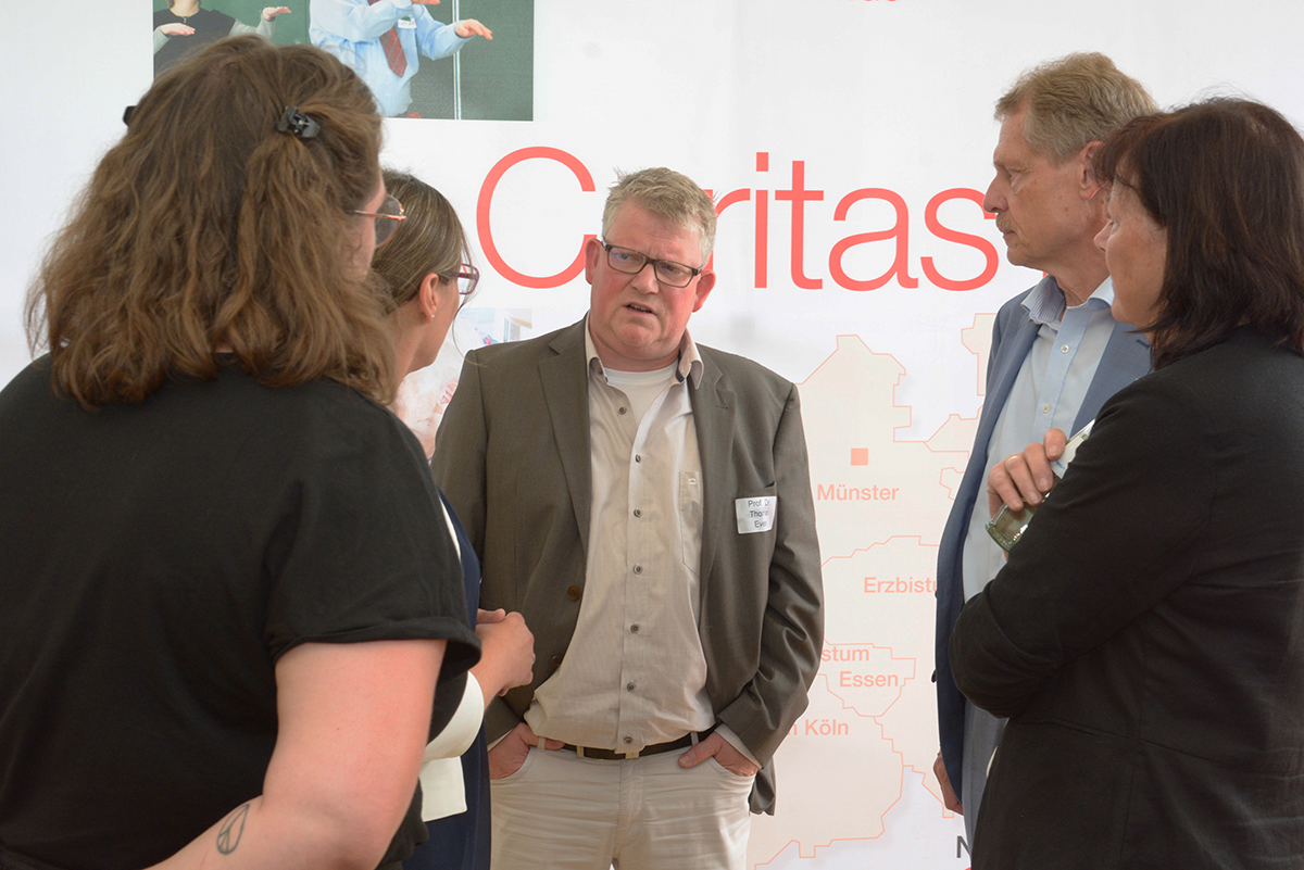 Prof. Dr. Thomas Evers steht vor einem Faltdisplay der Caritas in NRW und spricht mit vier Akteur/innen der CiNW-Fachtagung zur Umsetzung des Pflegeberufegesetzes in Essen (Foto: Caritas | Christoph Grätz)