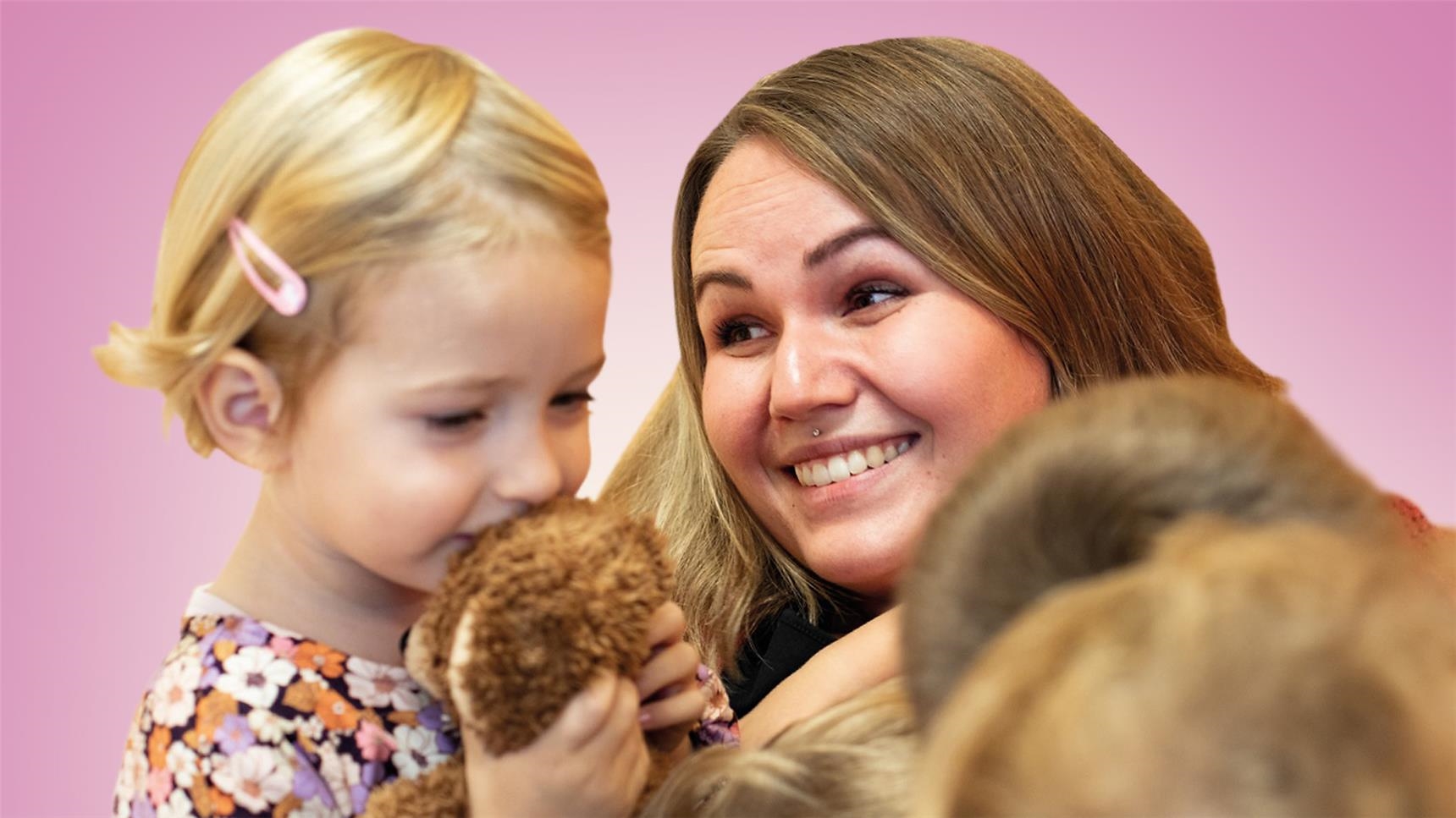 Eine junge Frau strahlt ein kleines Mädchen an, welches einen Teddybären in der Hand hält.