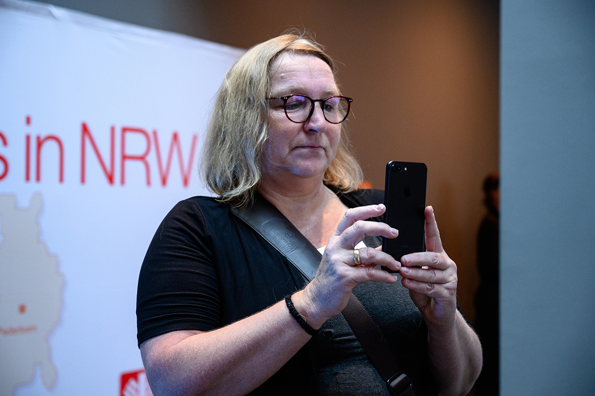 Sabine Depew steht vor einem Faltdisplay der Caritas in NRW und blickt dabei auf ein Smartphone (Charles Yunck)