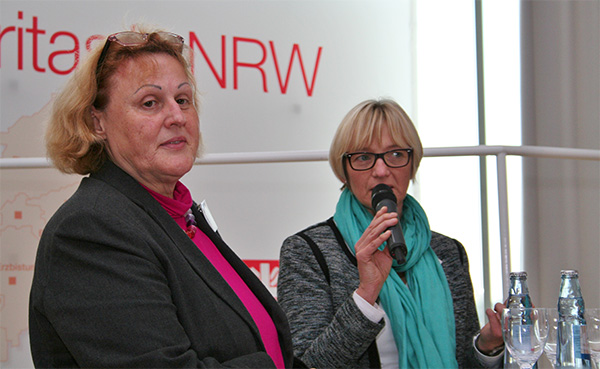 Zwei Rednerinnen stehen auf dem Podium, eine der beiden Frauen hält ein Mikrofon in der Hand und hält eine Rede. Im Hintergrund ist ein Faltdisplay der Caritas in NRW zu sehen. (Markus Lahrmann)